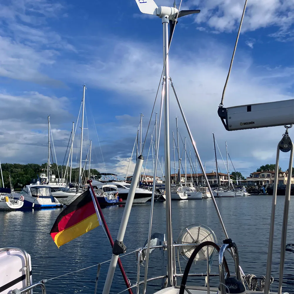 Windgenerator auf Edelstahl-Mast am Heck einer Segelyacht mit deutscher Flagge im Hafen bei blauem Himmel – zuverlässige Stromerzeugung durch Wind, ideal ergänzend zur Solaranlage für lange Liegezeiten und Ankernächte.