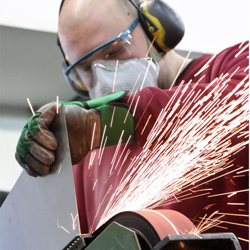 Metallarbeiter mit Schutzbrille und Handschuhen schleift eine Metallkante und erzeugt dabei Funkenflug.