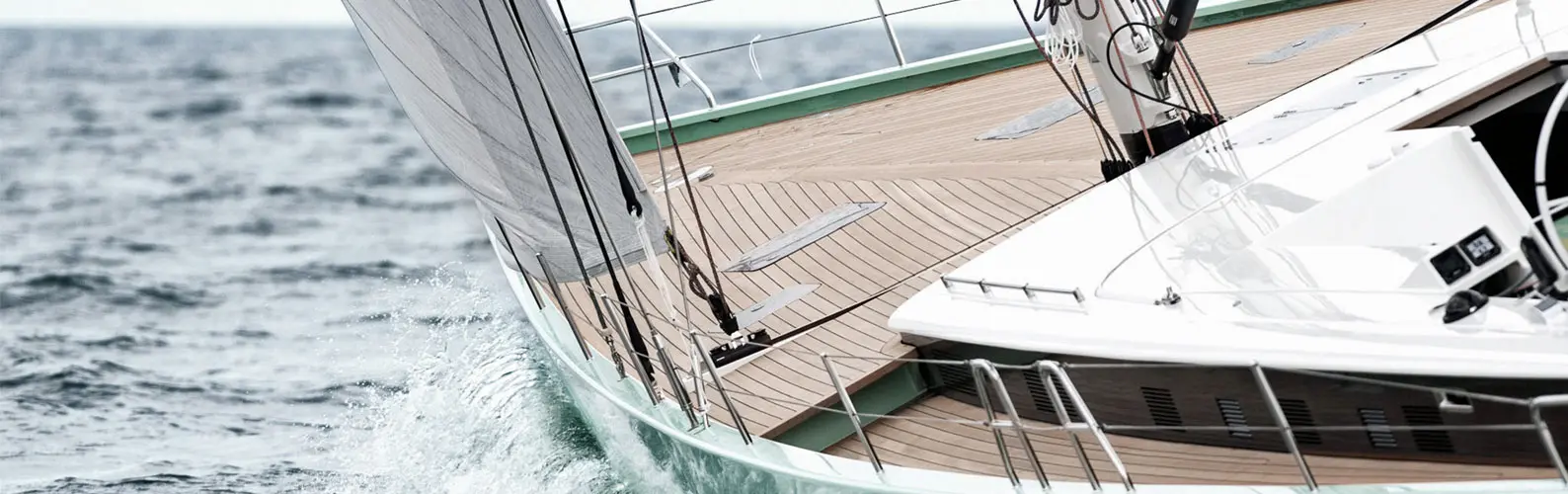 Luxusyacht mit Teakholzdeck segelt hart am Wind, im Vordergrund sind polierte Edelstahl Relingteile zu sehen